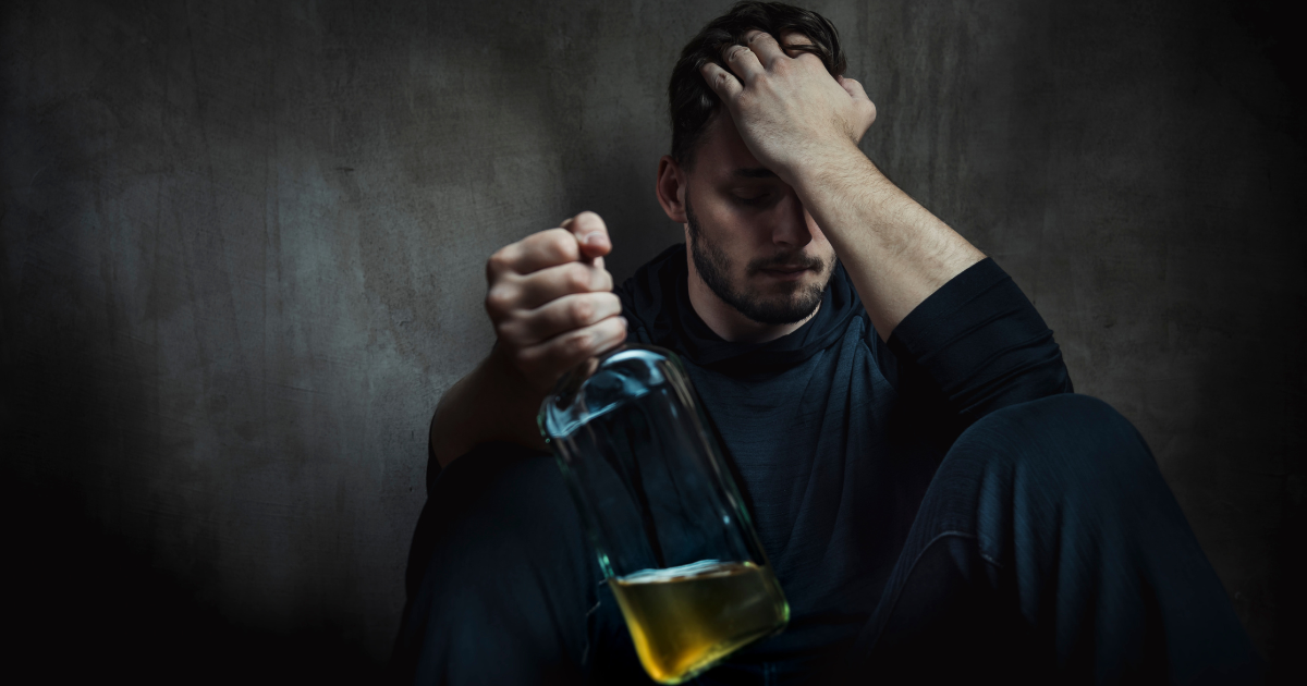 Sad looking man in black shirt sits against a wall holding a bottle of alcohol.