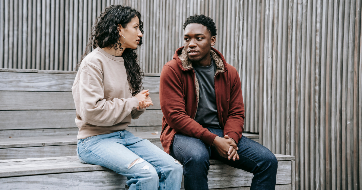 A woman and man wearing jeans and sweatshirts sit on a wooden bench having a serious conversation. 