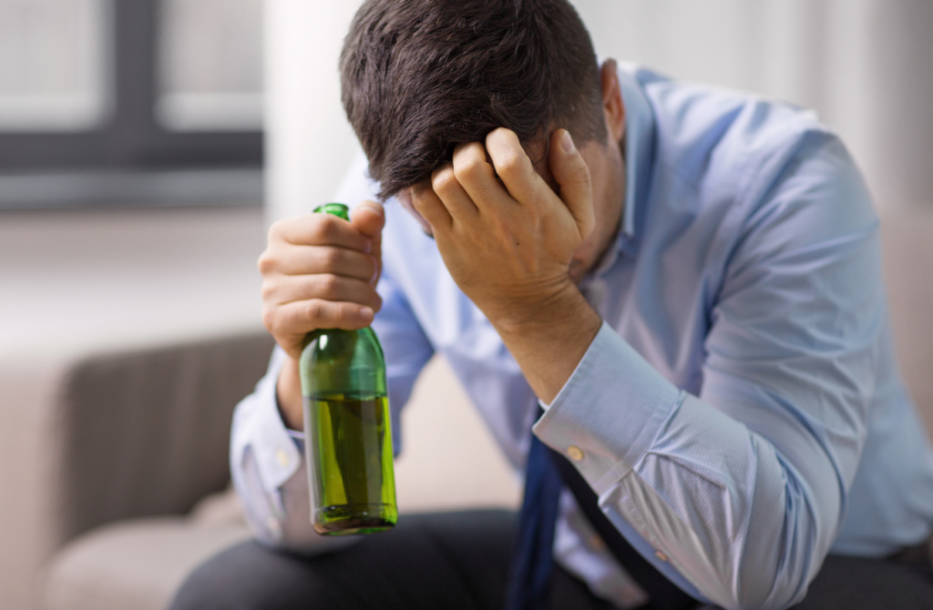 A man wearing a blue button-down shirt and tie clutches a bottle of beer and appears to be crying. 