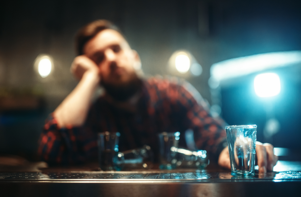 A man in a plaid shirt sits at a bar in front of empty shot glasses. He's got his hand to his head and looks sad. 