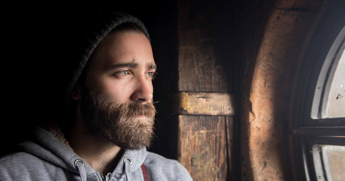 A man with a beard looks out a circular window with a wooden frame. He looks sad.
