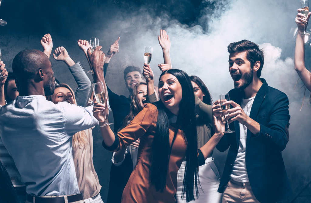 A group of people raise glasses of alcohol while dancing in a smoky room. 