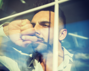 A bearded man in a collard shirt looks longingly through a window. He looks sad or upset, trying to find something. 
