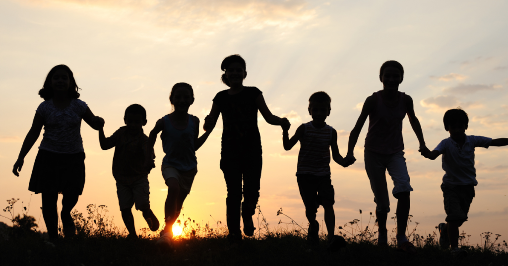 Seven children of various sizes holding hands are silhouetted against a sunset in a field.
