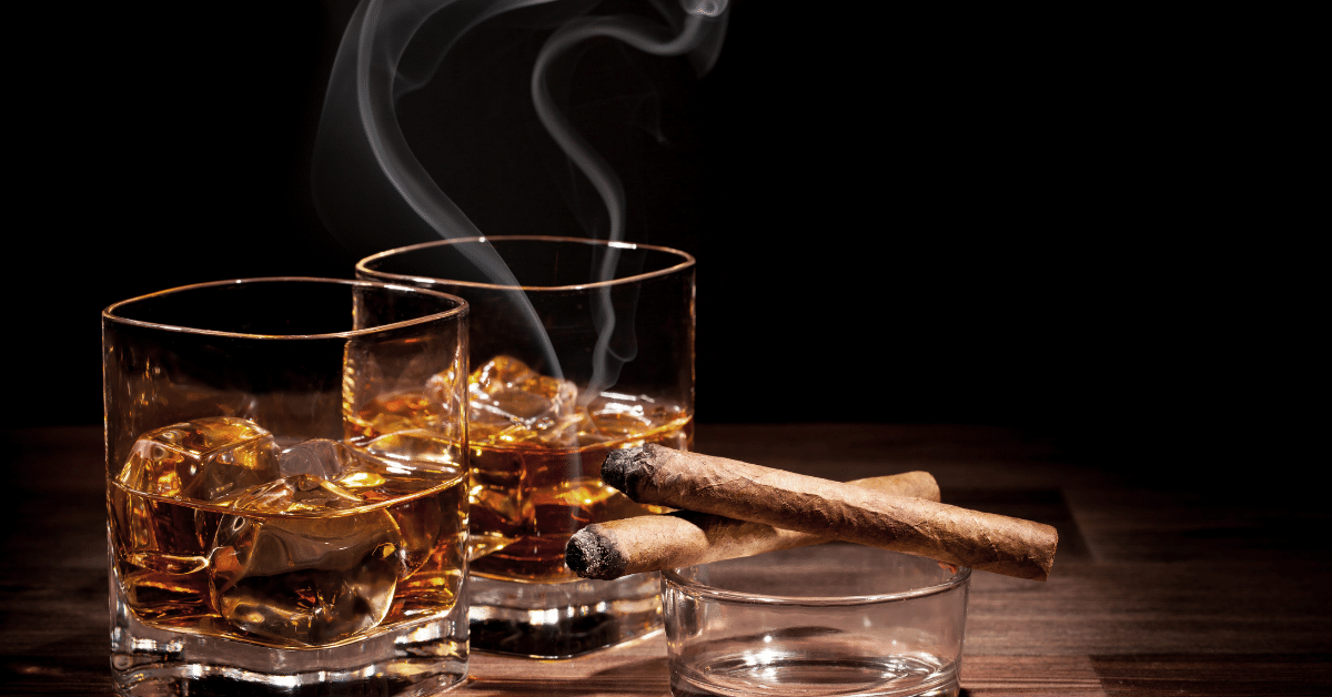 Whiskey in two glasses on a table in a dark room with a lit cigar balanced on empty glasses to signify familiar tastes and smells, which are triggers for those in recovery despite drinking beverages with no and low alcohol.